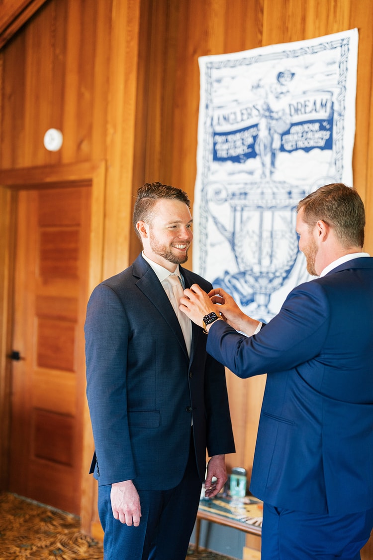 Groom smiling broadly as a groomsman pins his boutonniere in a warmly lit getting-ready suite.