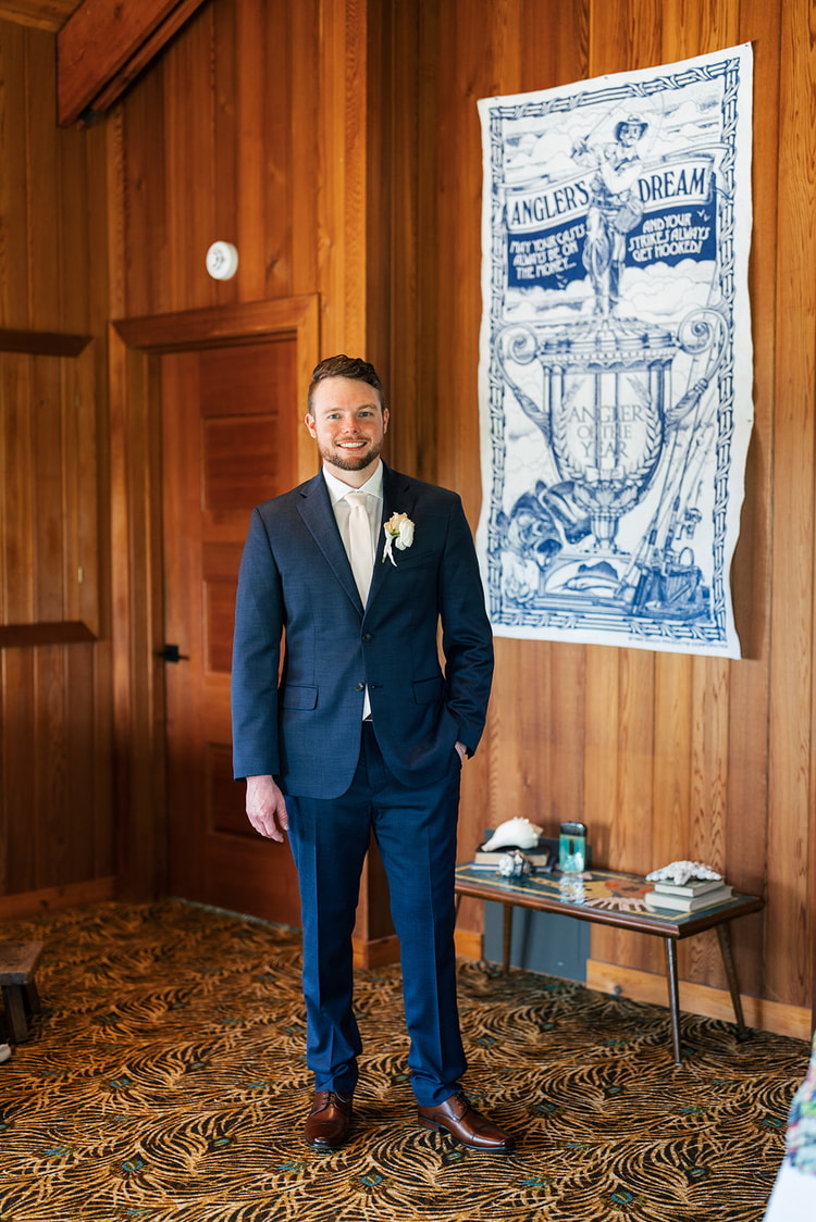 Groom standing confidently in a navy suit with a white boutonniere inside a wood-paneled room.