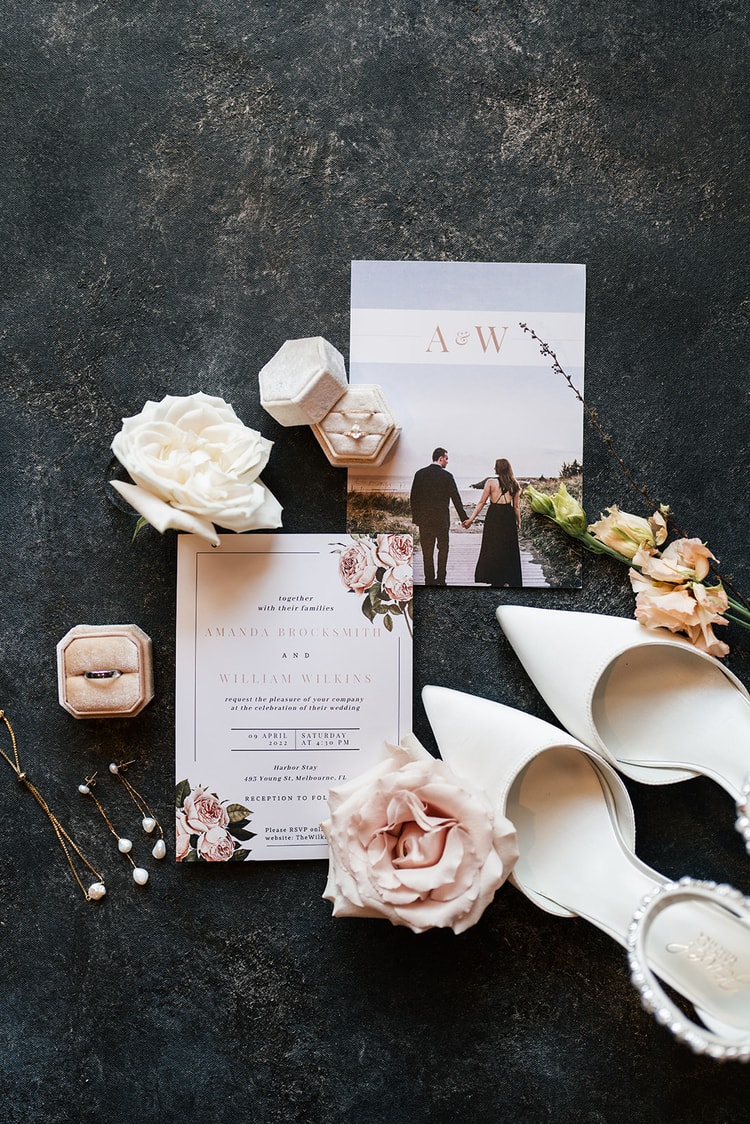 Flat-lay wedding details: floral invitation suite, ring box, pearl drop earrings, white heels, and blush and ivory roses on a dark stone surface.