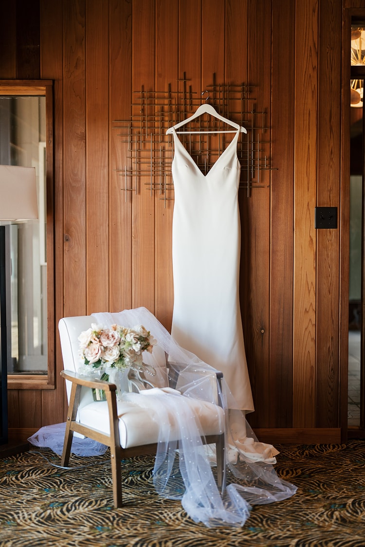 White wedding gown on a hanger against wood paneling, veil and bridal bouquet resting on a chair below.