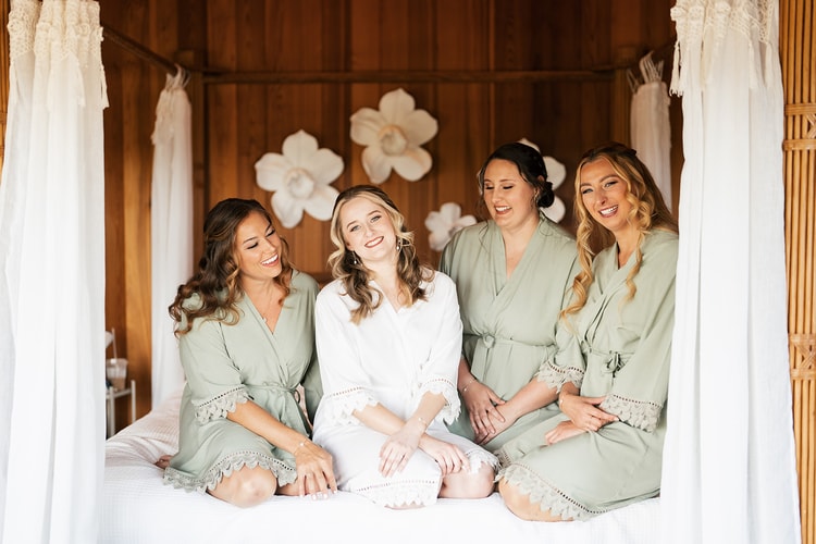 Bride in a white robe laughs with three bridesmaids in sage green robes while seated together on a canopy bed.