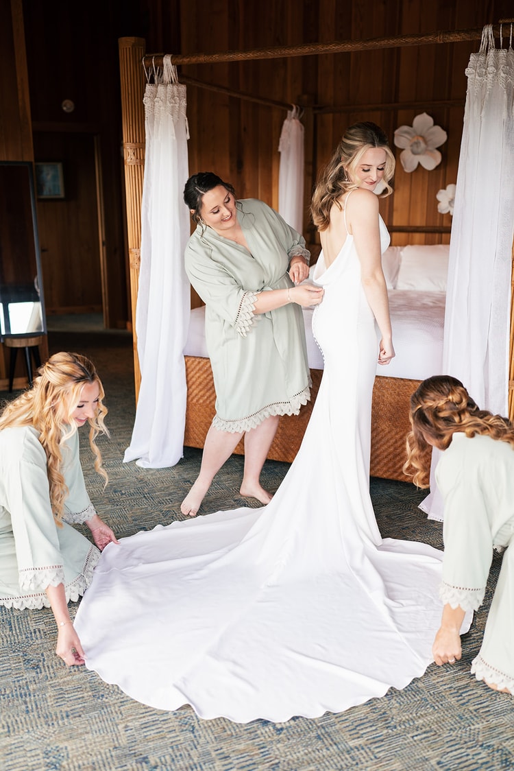 Bridesmaids in sage robes help the bride into her wedding gown, arranging the long train across the floor.