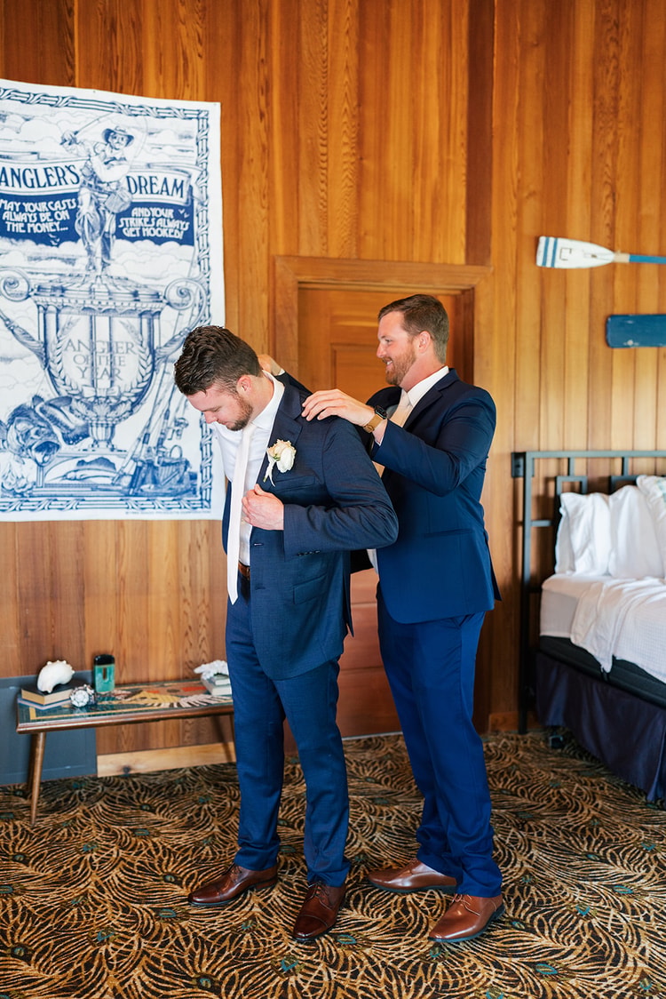 Groomsman in a navy suit helps the groom adjust his boutonniere while both smile in a wood-paneled room.