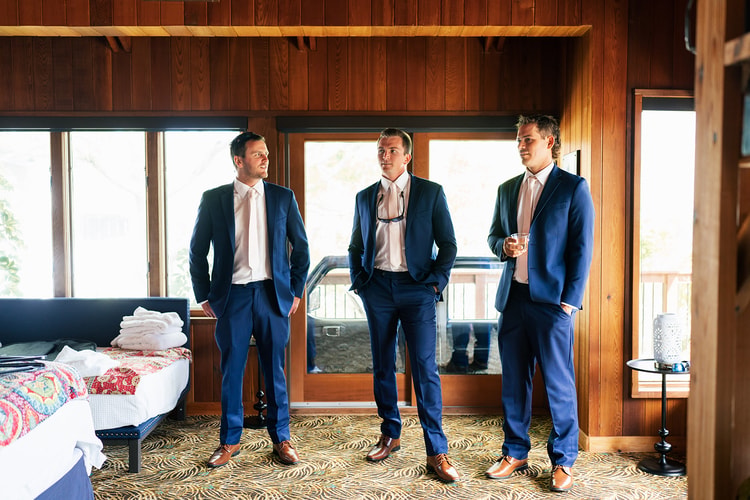 Three groomsmen in matching navy suits standing and chatting in a sun-filled room before the ceremony.
