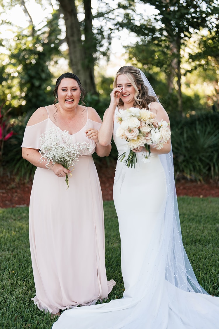 Bride and bridesmaid standing side by side on the lawn; the bride wipes a tear while her bridesmaid beams beside her.
