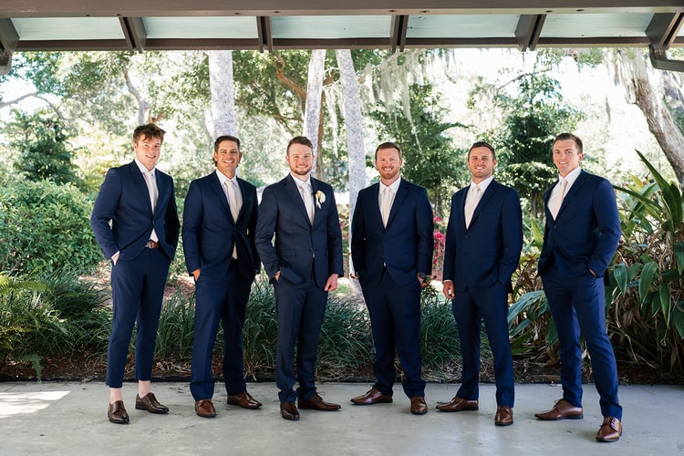 Six groomsmen in navy suits posed in a row under a covered pavilion surrounded by tropical greenery.