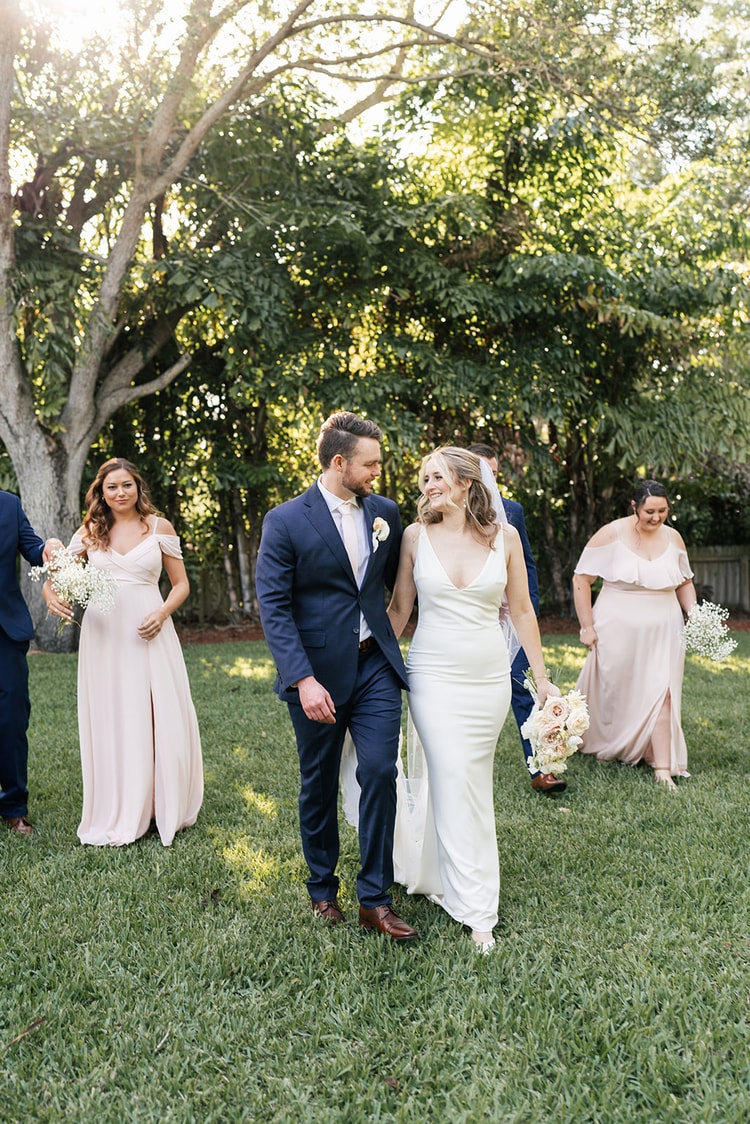 Bride and groom walk together across the grass, bridesmaids strolling behind them in the dappled afternoon light.