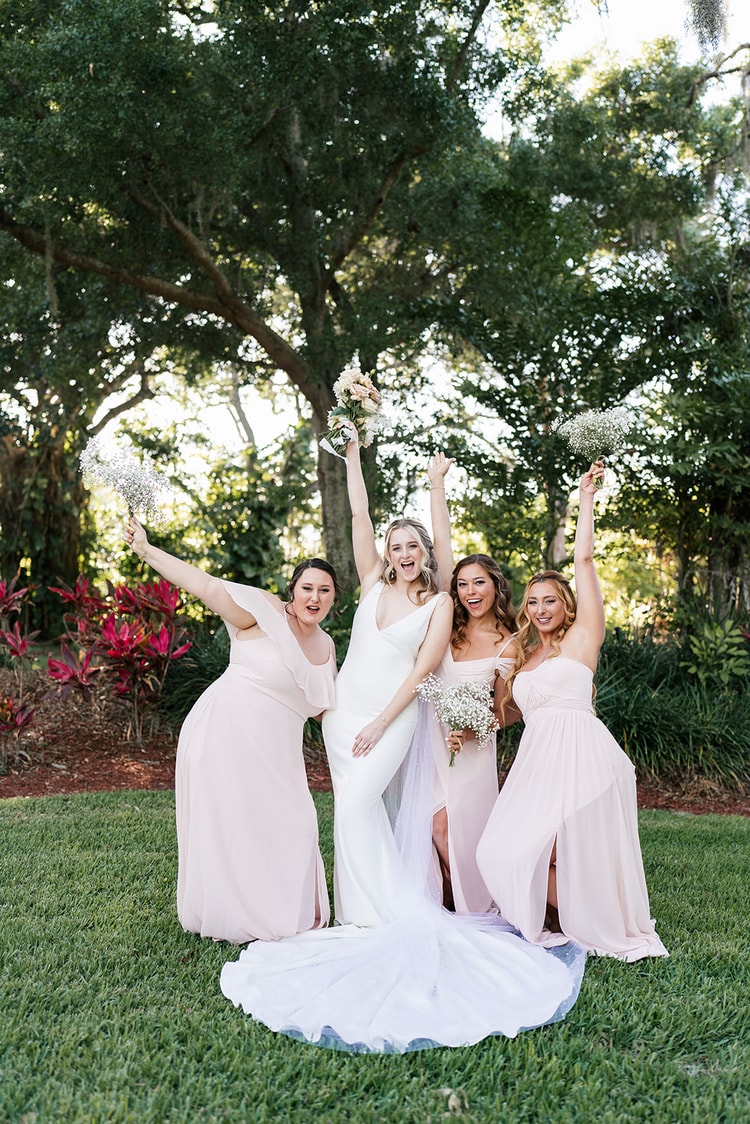 Bride and three bridesmaids in blush gowns celebrate on the lawn with arms raised, laughing joyfully.