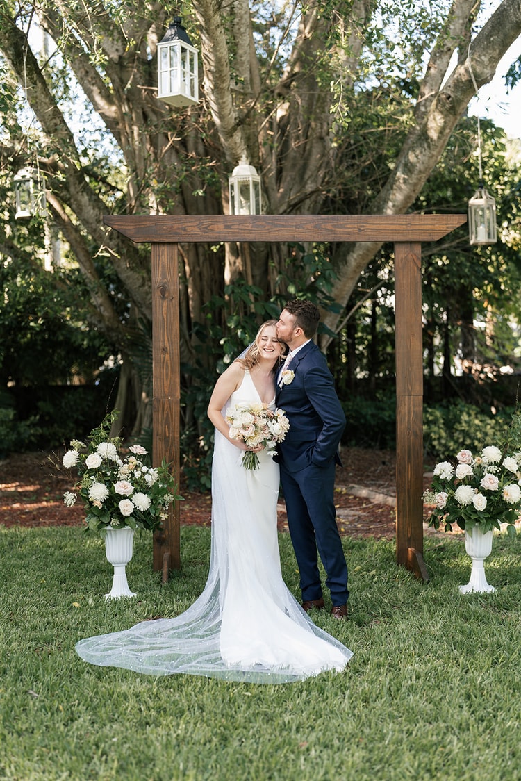 Couple posing at the ceremony arch after the wedding, white floral urns and hanging lanterns surrounding them.