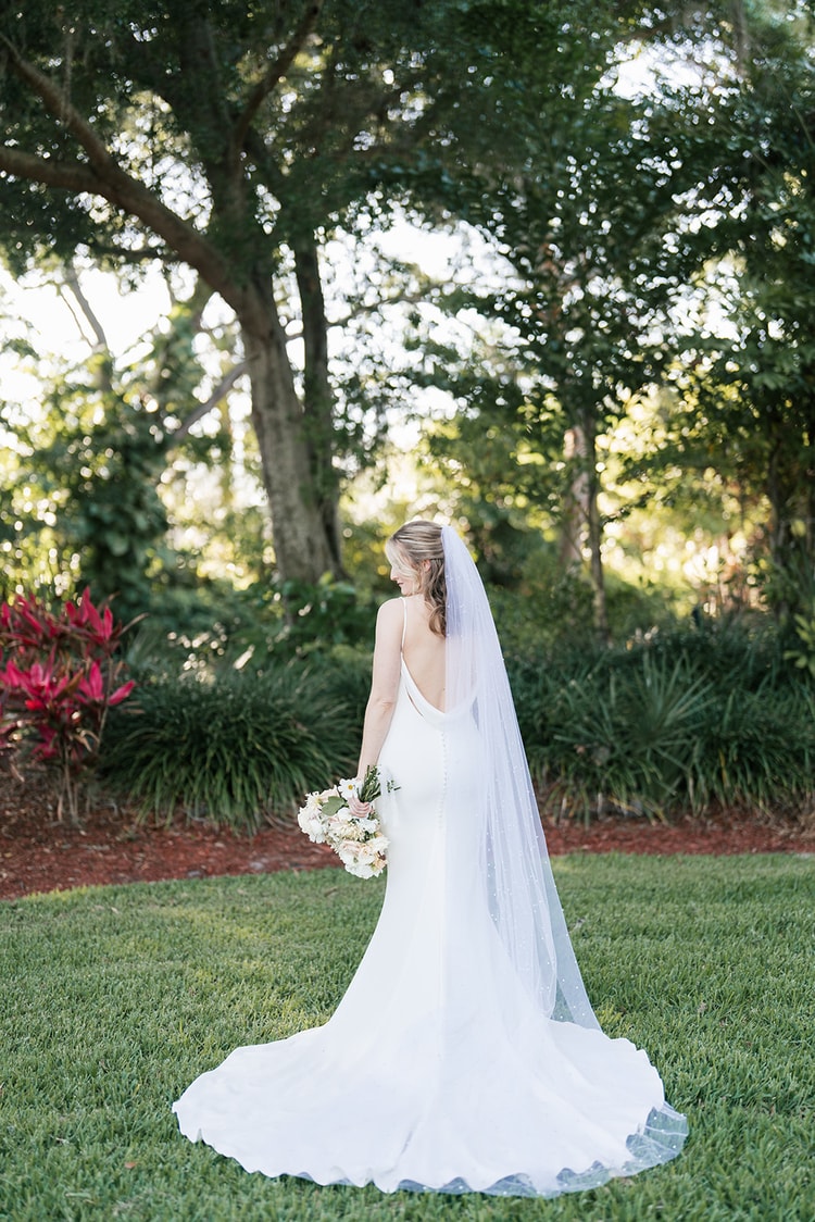 Bride seen from behind in her open-back gown and cathedral veil, holding a soft blush bouquet amid lush garden greenery.