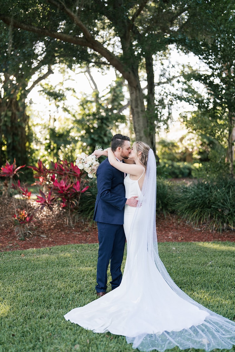 Bride and groom share a close kiss in a lush garden, the bride's cathedral veil pooling around them.