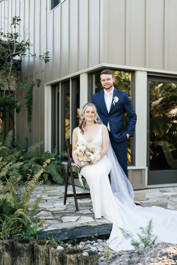 Bride seated on a folding chair holding her bouquet, groom standing behind her, both smiling at the camera in a garden courtyard.
