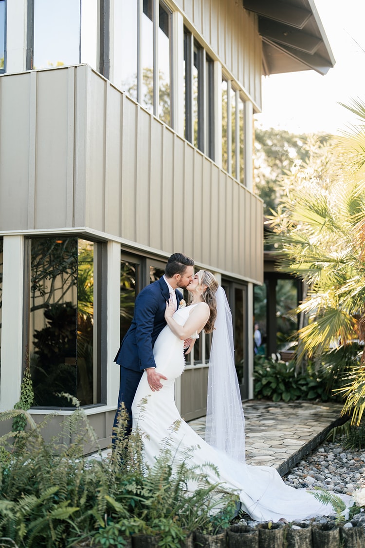 Couple sharing a golden-hour kiss outside the venue, tropical palms framing the scene.