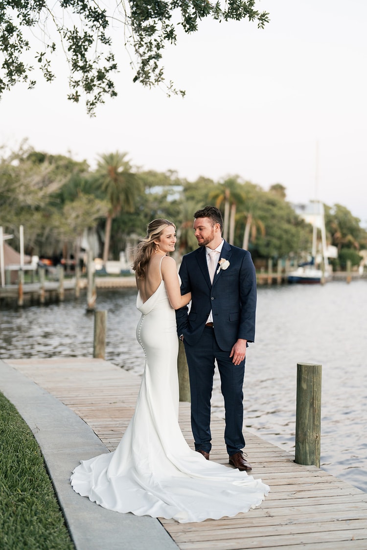 Bride glances back at the groom while they stand together on a wooden dock, her train trailing behind her at sunset.