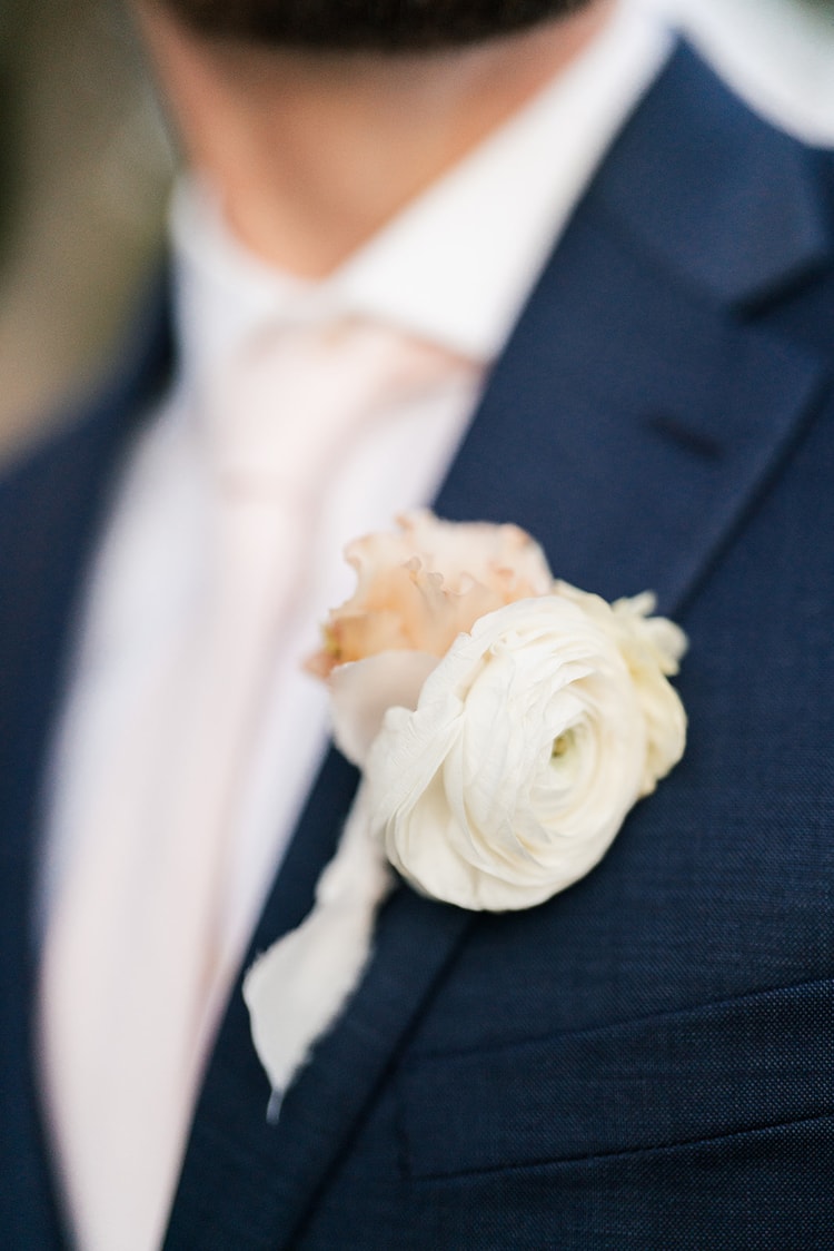Close detail of the groom's boutonniere: a white ranunculus and blush bud pinned to a navy lapel.