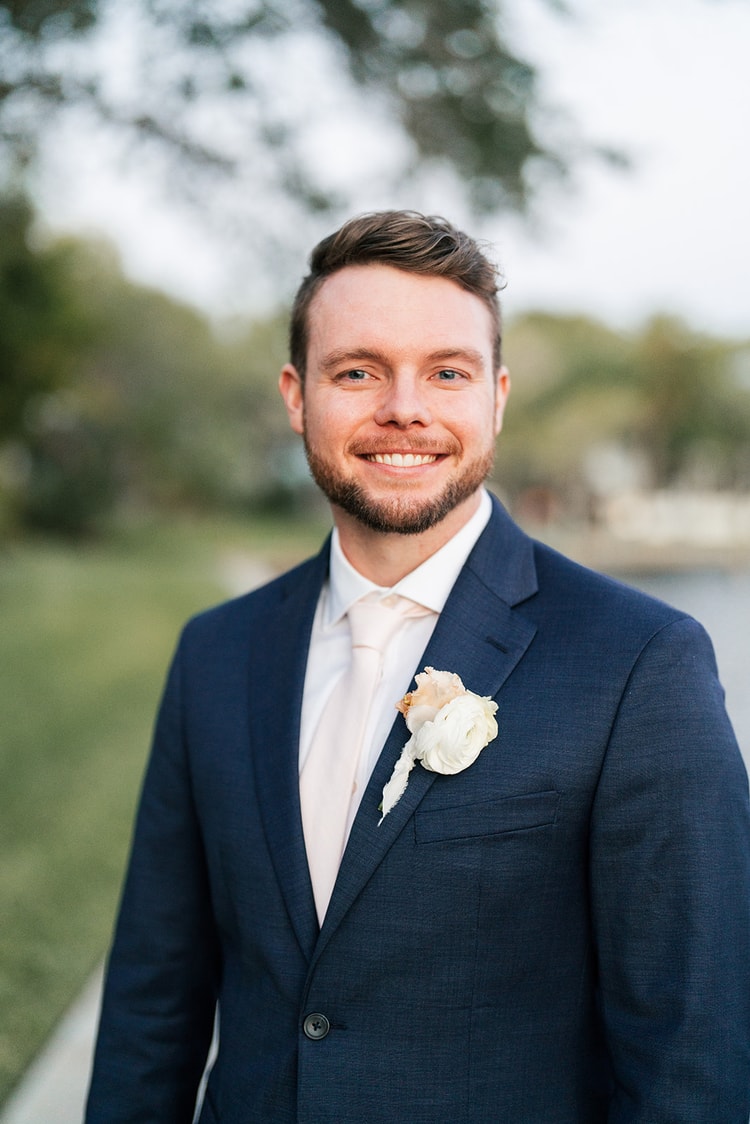 Groom portrait in a navy suit with a white ranunculus boutonniere, smiling outdoors near the water.