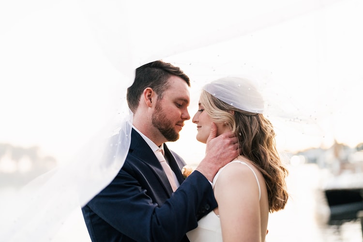 Couple under the bride's flowing veil, faces nearly touching in a quiet, intimate moment on the waterfront dock.