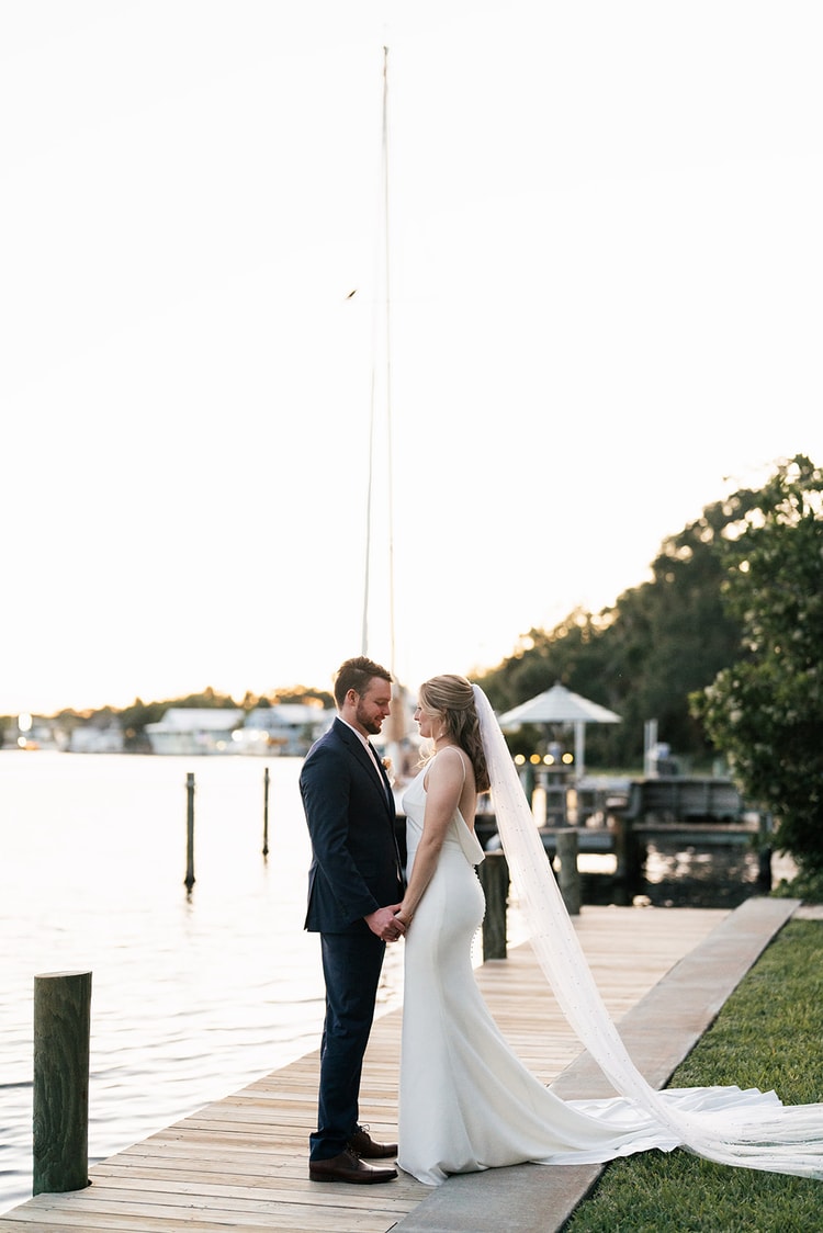 Couple standing hand in hand at golden hour on a dock, sailboat mast rising between them against a pale sky.