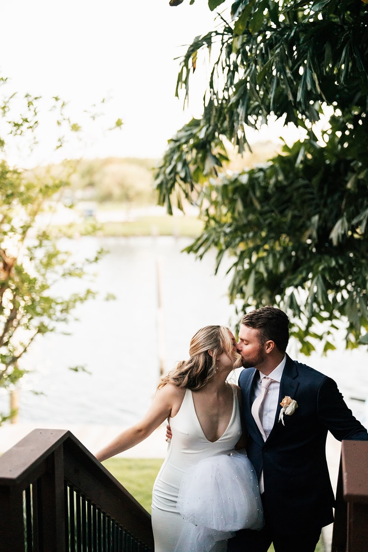 Newlyweds kissing on a balcony staircase with a view of the sparkling waterway through tropical foliage.
