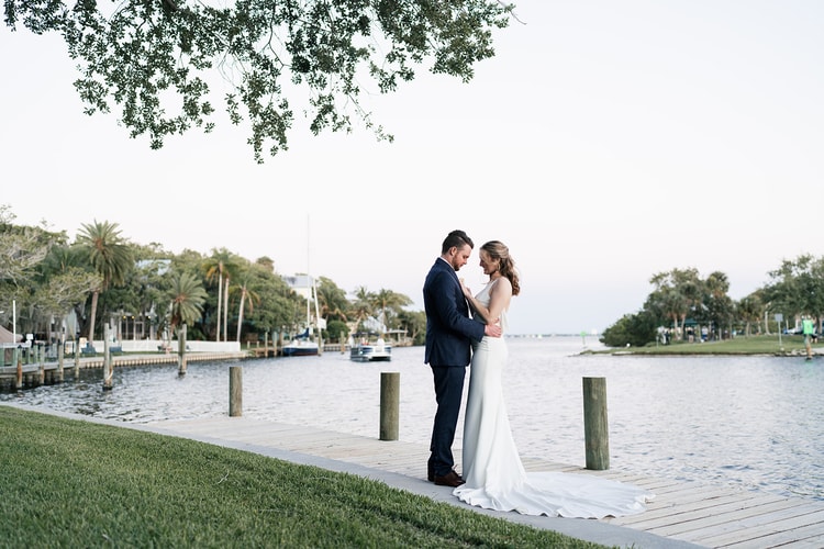 Bride and groom standing forehead to forehead on a waterfront dock at dusk, the calm river stretching behind them.