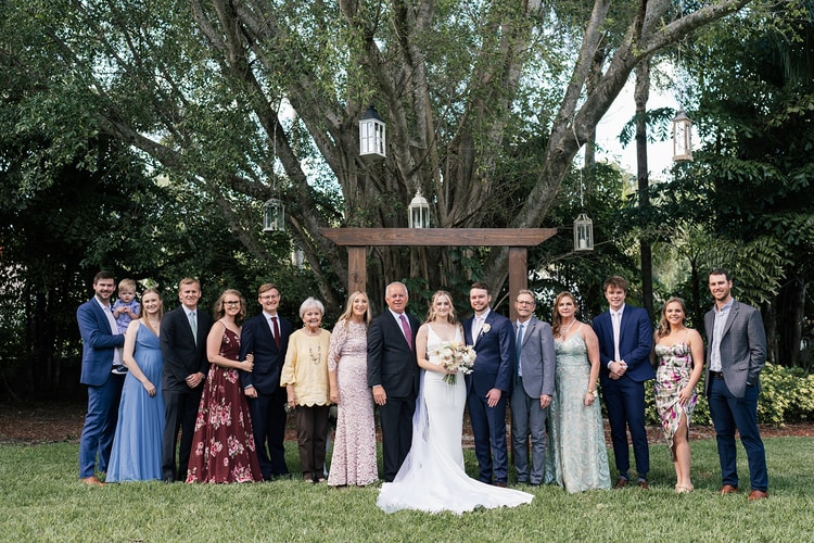 Extended family group portrait in front of a rustic wooden arch with lanterns hanging from a sprawling tree.