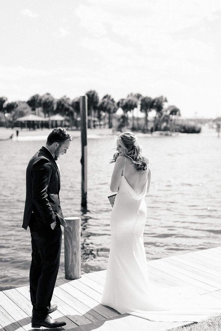 Black-and-white first-look moment on a waterfront dock; groom looks down at his vows while the bride laughs with her hand over her mouth.