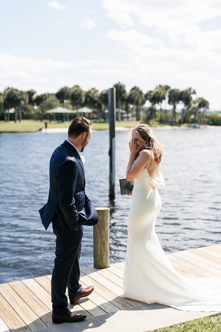 First-look on the dock: bride covering her face with delight as the groom turns to see her for the first time, water shimmering behind them.