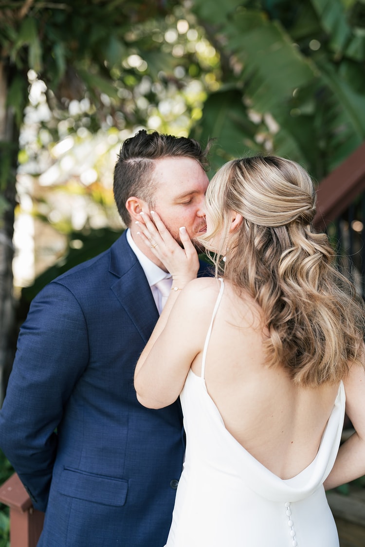 Bride cups the groom's face and kisses him tenderly amid lush tropical foliage.