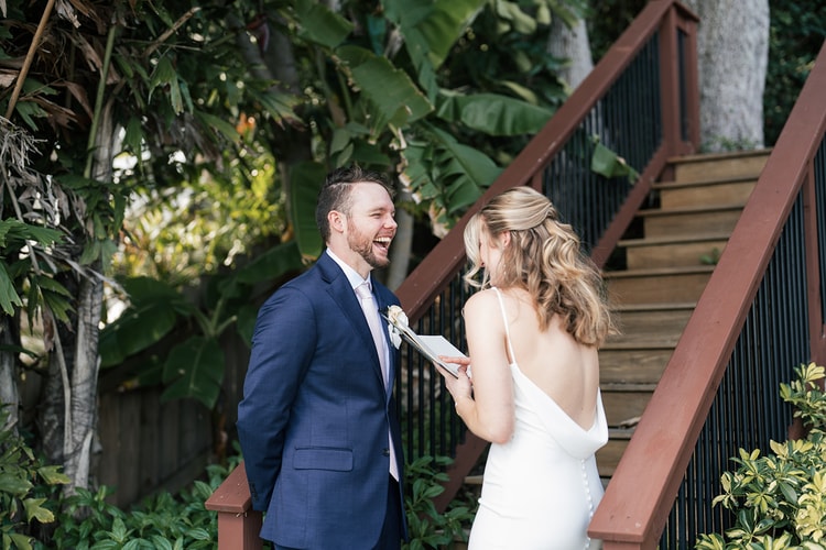 Groom laughing out loud as the bride reads from a vow book during their private first-look exchange beside garden stairs.