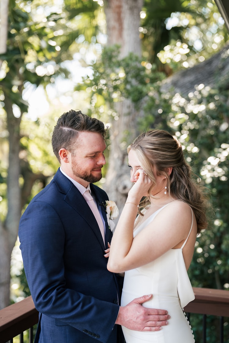 Groom gently holds the bride's face as she wipes a happy tear during an emotional moment on a wooden deck.