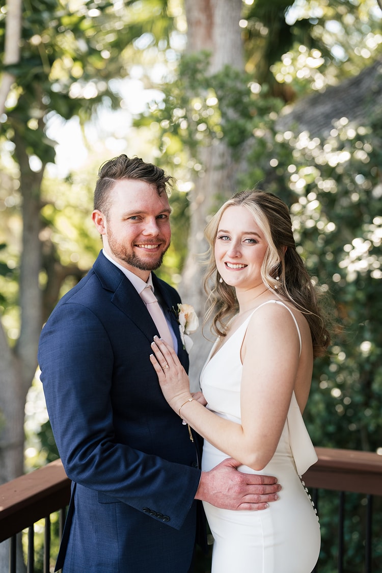Bride and groom smiling together on a deck railing framed by green trees, the groom's arm around her waist.