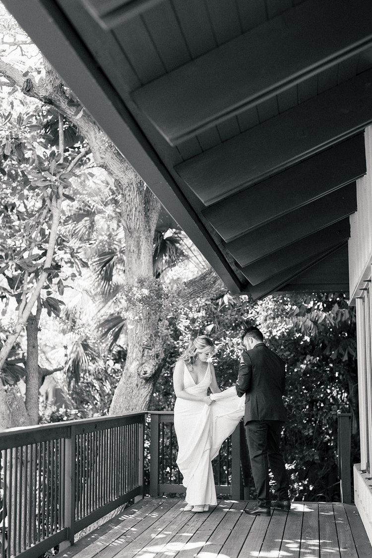 Black-and-white portrait of the couple standing close on a covered deck, surrounded by leafy trees.
