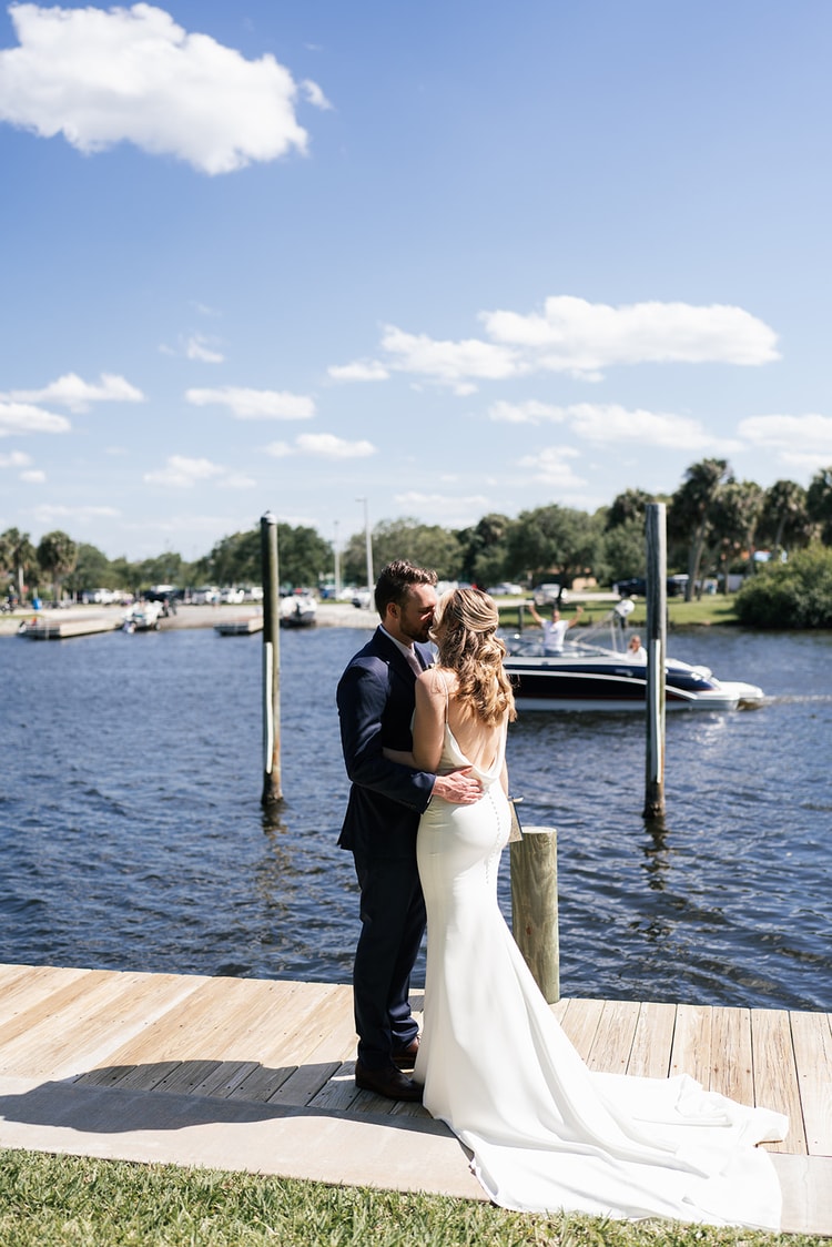 Couple sharing a kiss on a waterfront dock, the bride's long train spread across the wooden planks.