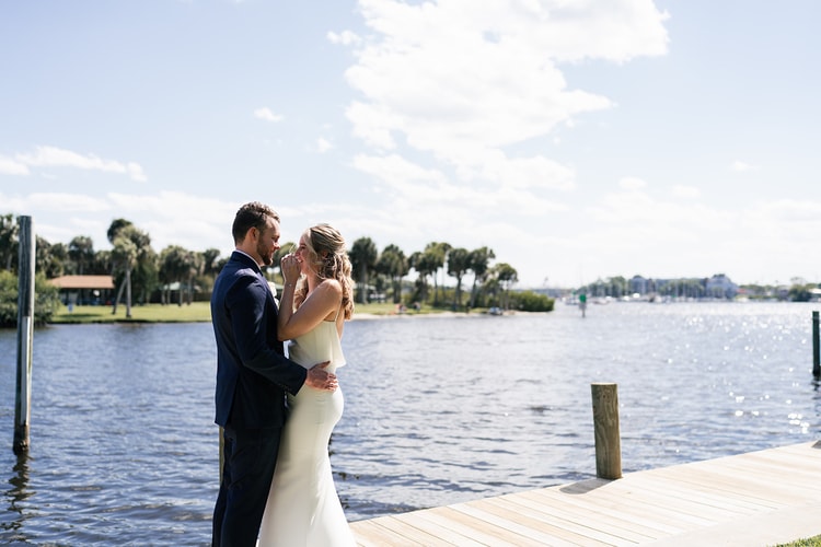 Bride and groom embrace on a sunny dock overlooking the water, the bride smiling up at him.