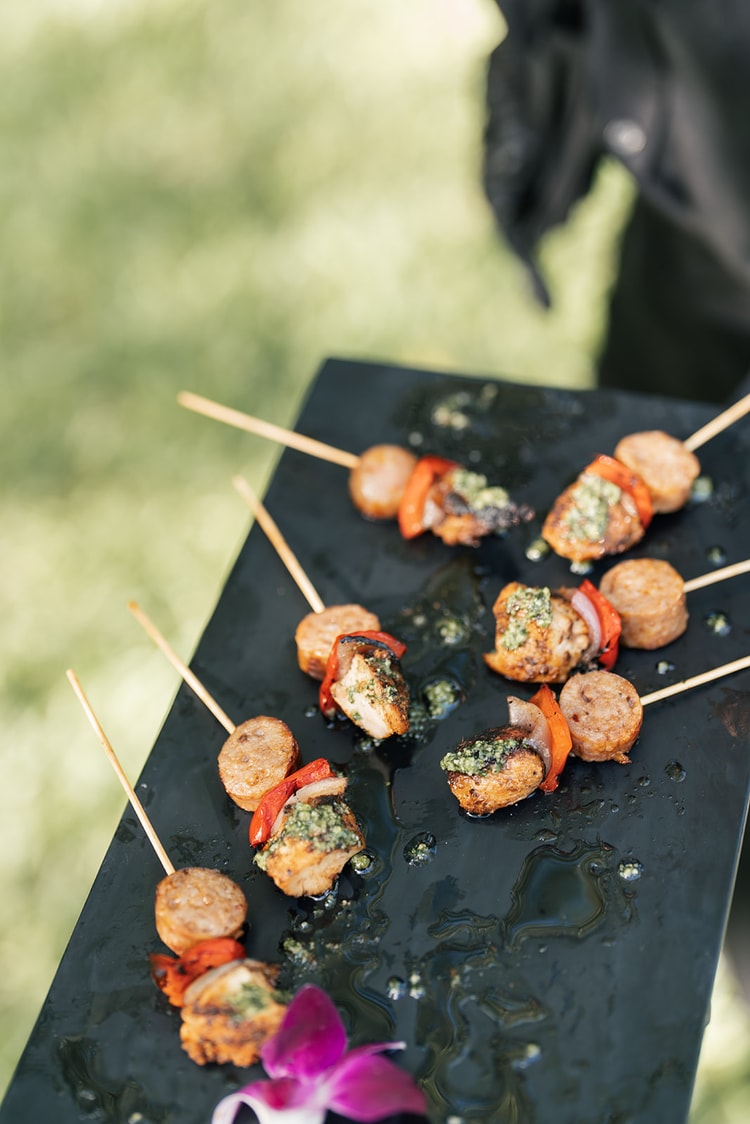 Cocktail-hour appetizer skewers with chicken, sausage, and roasted peppers drizzled with herb sauce on a slate tray.