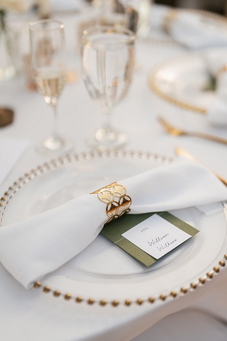 Reception place setting detail: white napkin with a gold napkin ring and a calligraphy name card on a beaded charger plate.