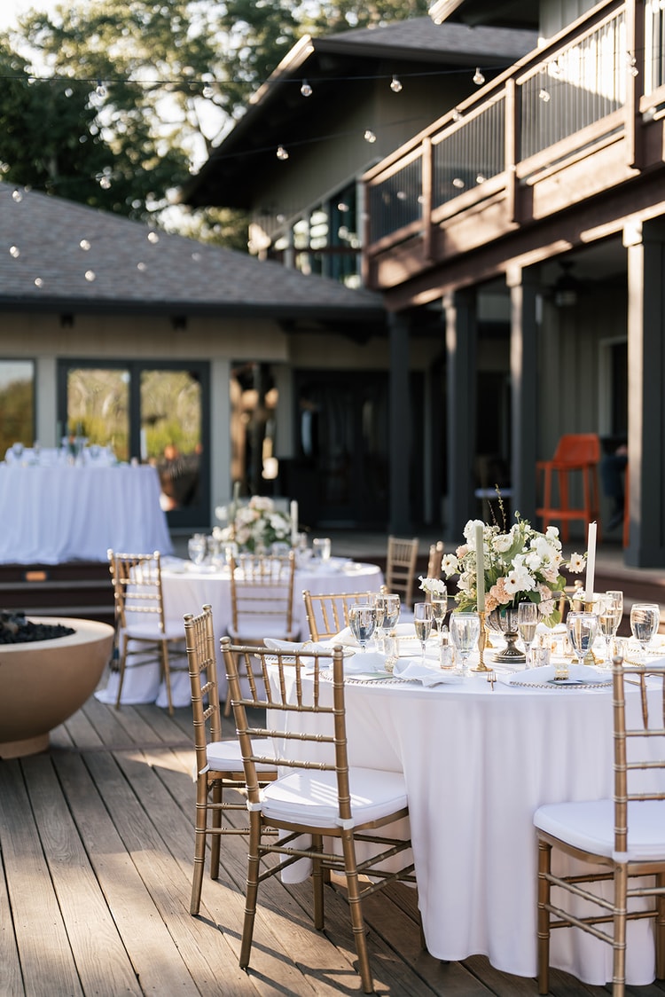 Outdoor reception setup on a deck: round tables with white linens, gold chiavari chairs, and a fire bowl feature.