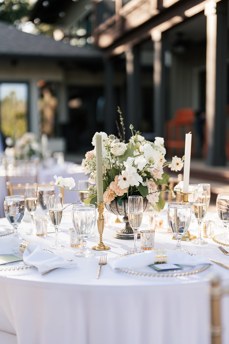 Lush blush and ivory floral centerpiece with sage-green taper candles on a white-linen reception table.