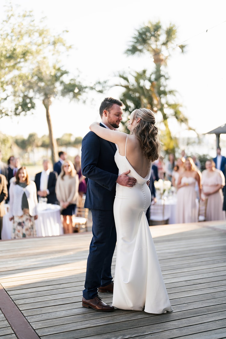 Couple sharing their first dance as newlyweds under golden-hour light on the outdoor deck, surrounded by watching guests.