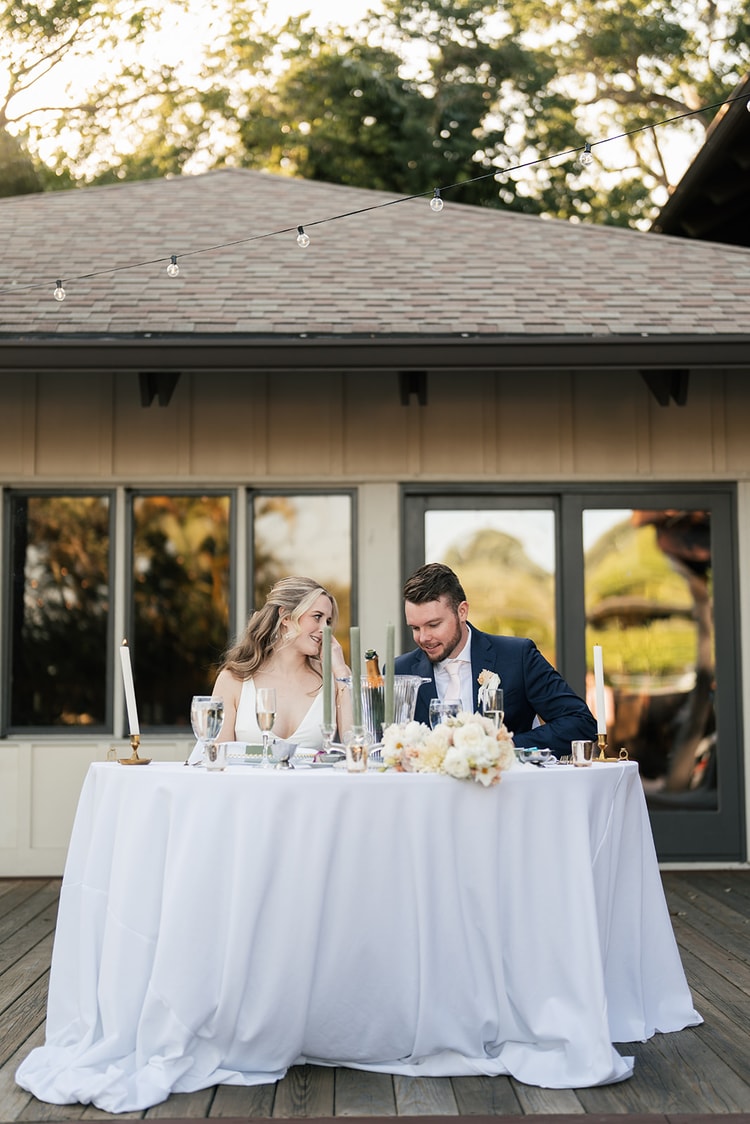Bride and groom seated together at the candlelit sweetheart table, sharing a quiet smile under string lights.