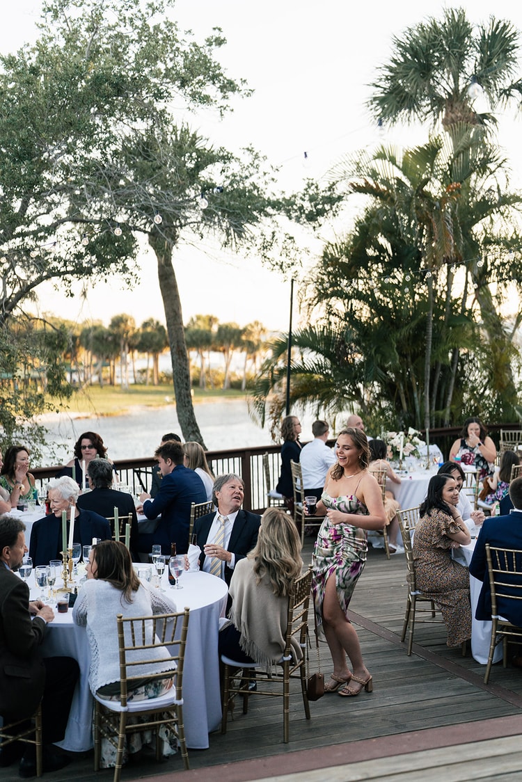 Reception guests laughing and conversing at their tables on the outdoor deck under string lights at dusk.