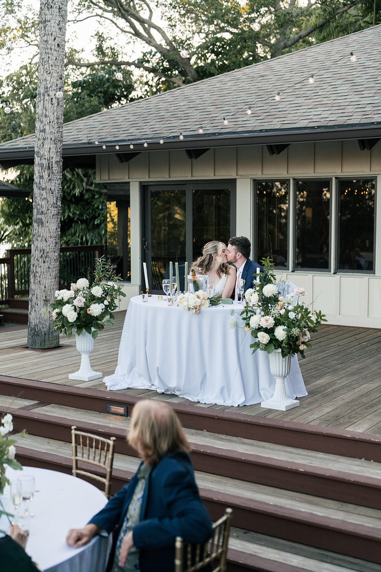 Bride and groom kissing at the sweetheart table during the reception, flower-filled urns framing them on either side.
