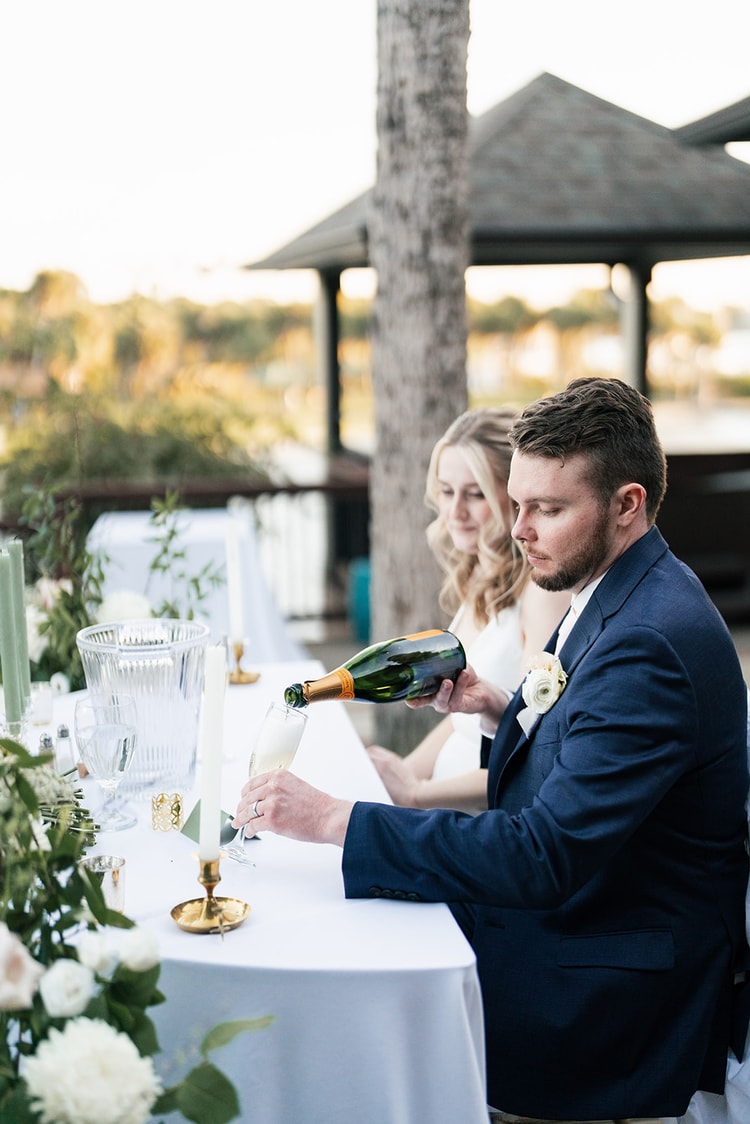 Groom pours champagne from a bottle at the sweetheart table as the bride looks on, floral arrangements glowing in the warm light.