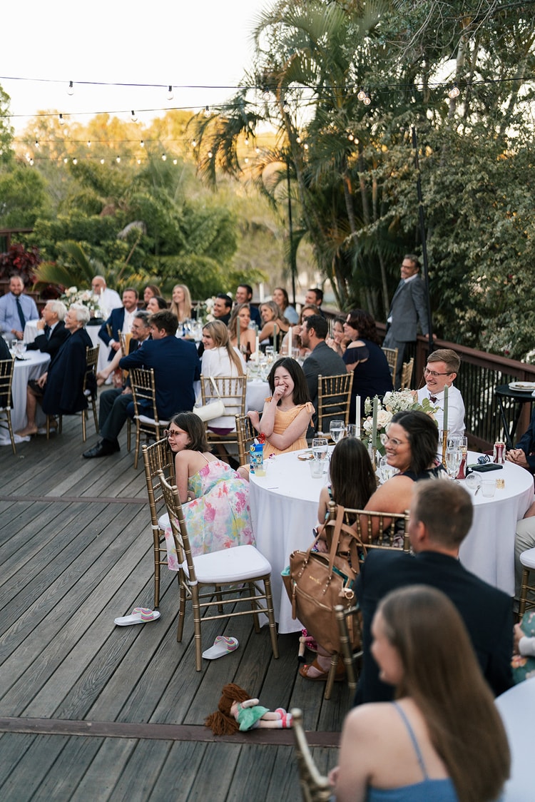 Guests seated across the outdoor reception deck, laughing during the speeches, string lights glowing overhead.