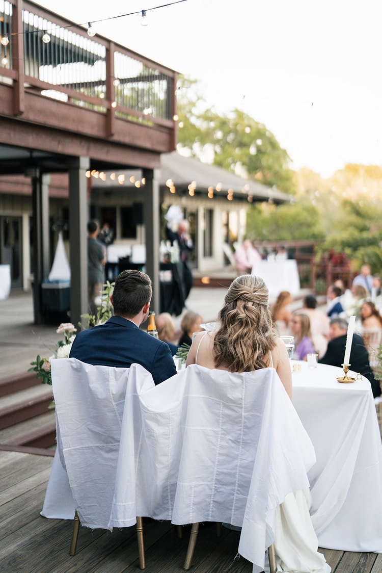 Bride and groom seen from behind at the sweetheart table, watching the reception activities unfold on the deck.