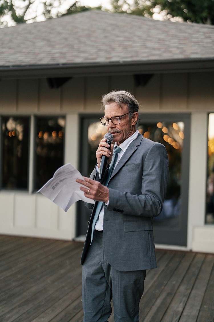 An older gentleman in a grey suit delivers a heartfelt wedding speech from the outdoor deck stage.