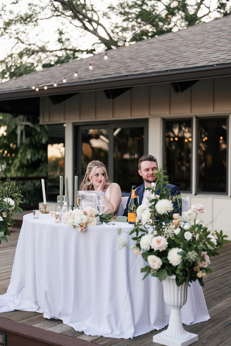 Couple at the sweetheart table listening to toasts, bride emotional as she watches, surrounded by white floral arrangements.