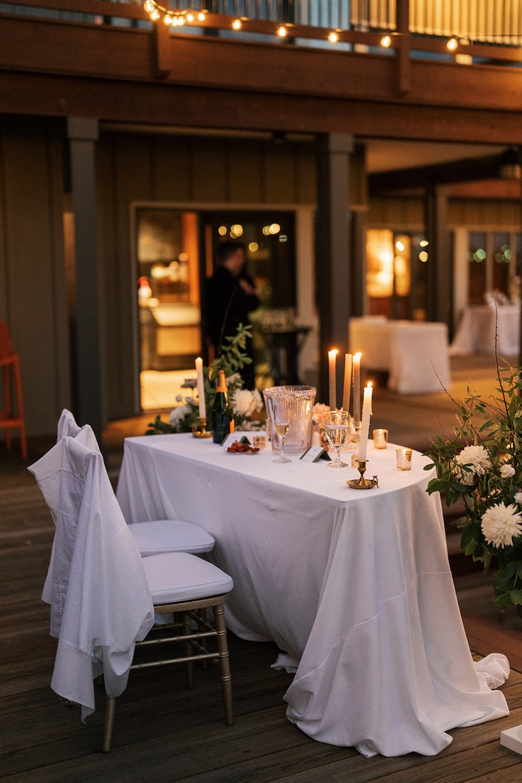 Candlelit sweetheart table on an outdoor deck at dusk, flanked by white dahlia arrangements and string lights overhead.