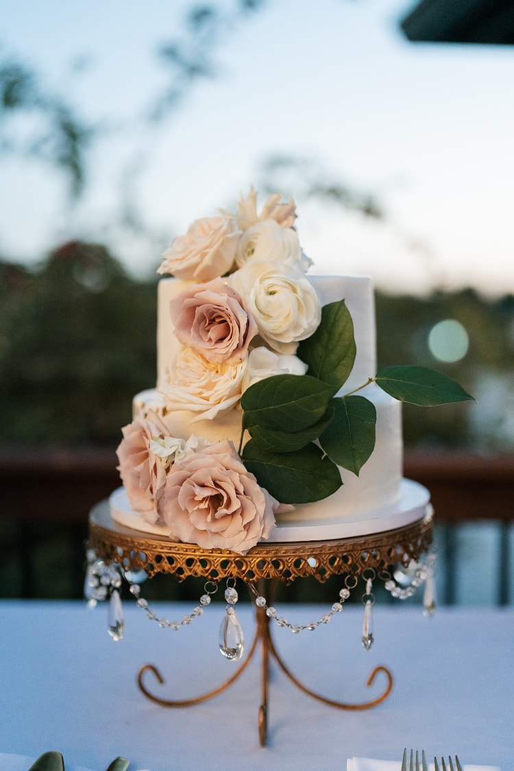 Two-tier white wedding cake adorned with cascading blush garden roses and ranunculus on a gold chandelier cake stand.