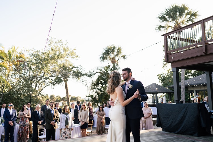 Couple shares their first dance on an outdoor wooden deck at golden hour, guests circled around them watching.